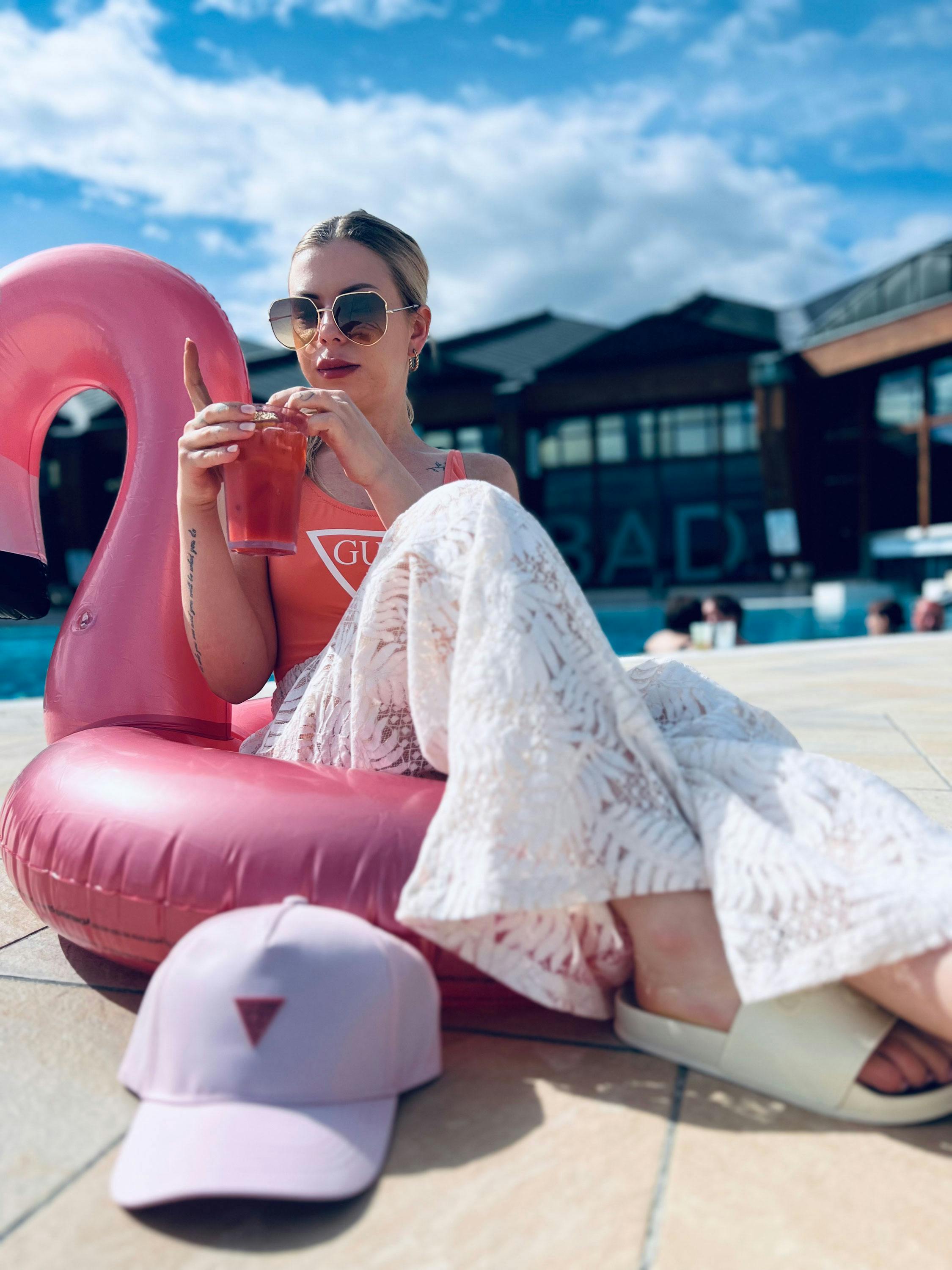 Person sitzt auf einem aufblasbaren pinkfarbenen Flamingo, trägt weiße Hose mit Muster und weiße Sandalen, hält ein Getränk in der Hand, vor ihr liegt eine hellrosa Kappe mit rotem Dreieck-Logo.