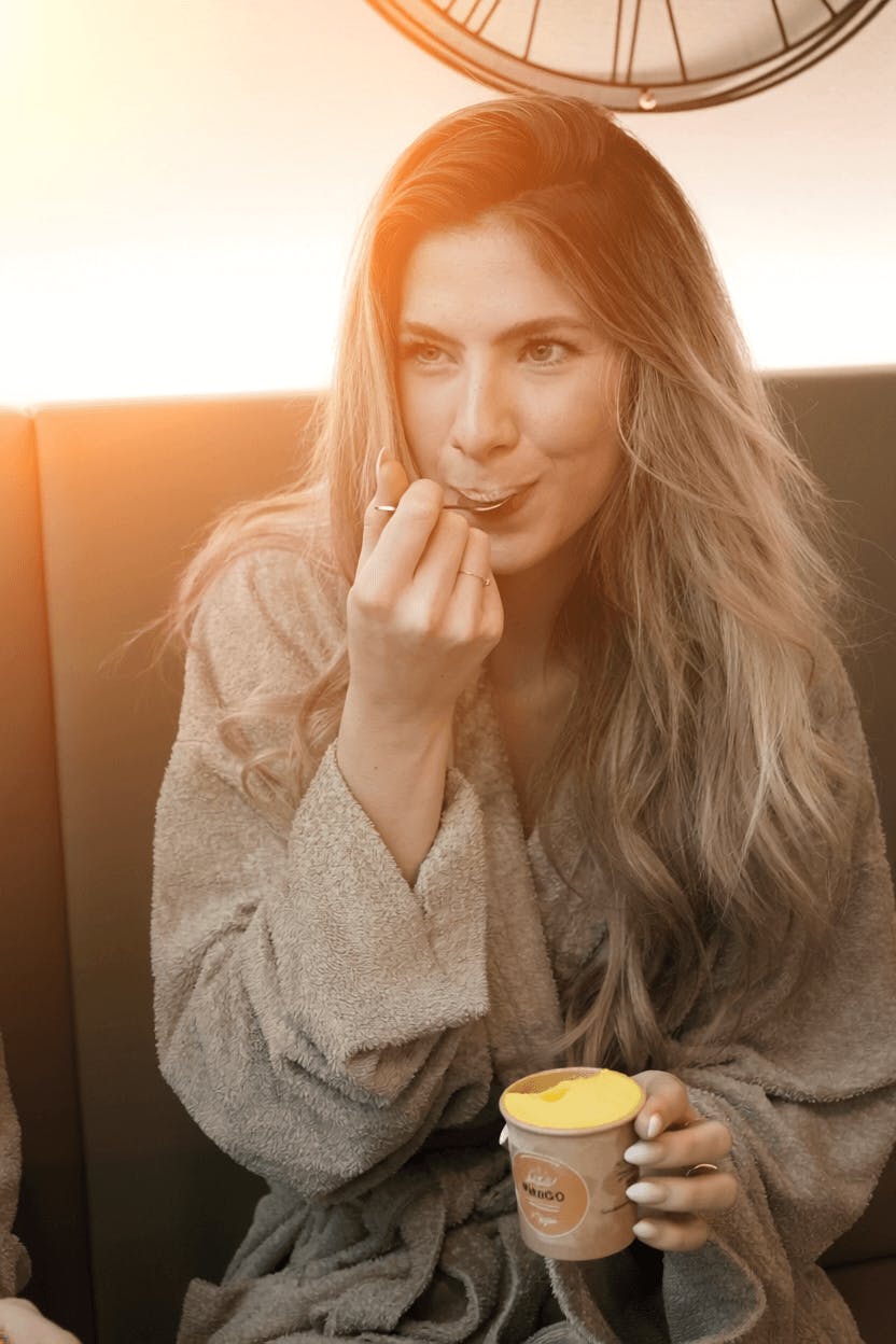 Frau mit langem, gewelltem Haar in Bademantel sitzt auf Sofa und hält Becher mit Eiscreme in der Hand