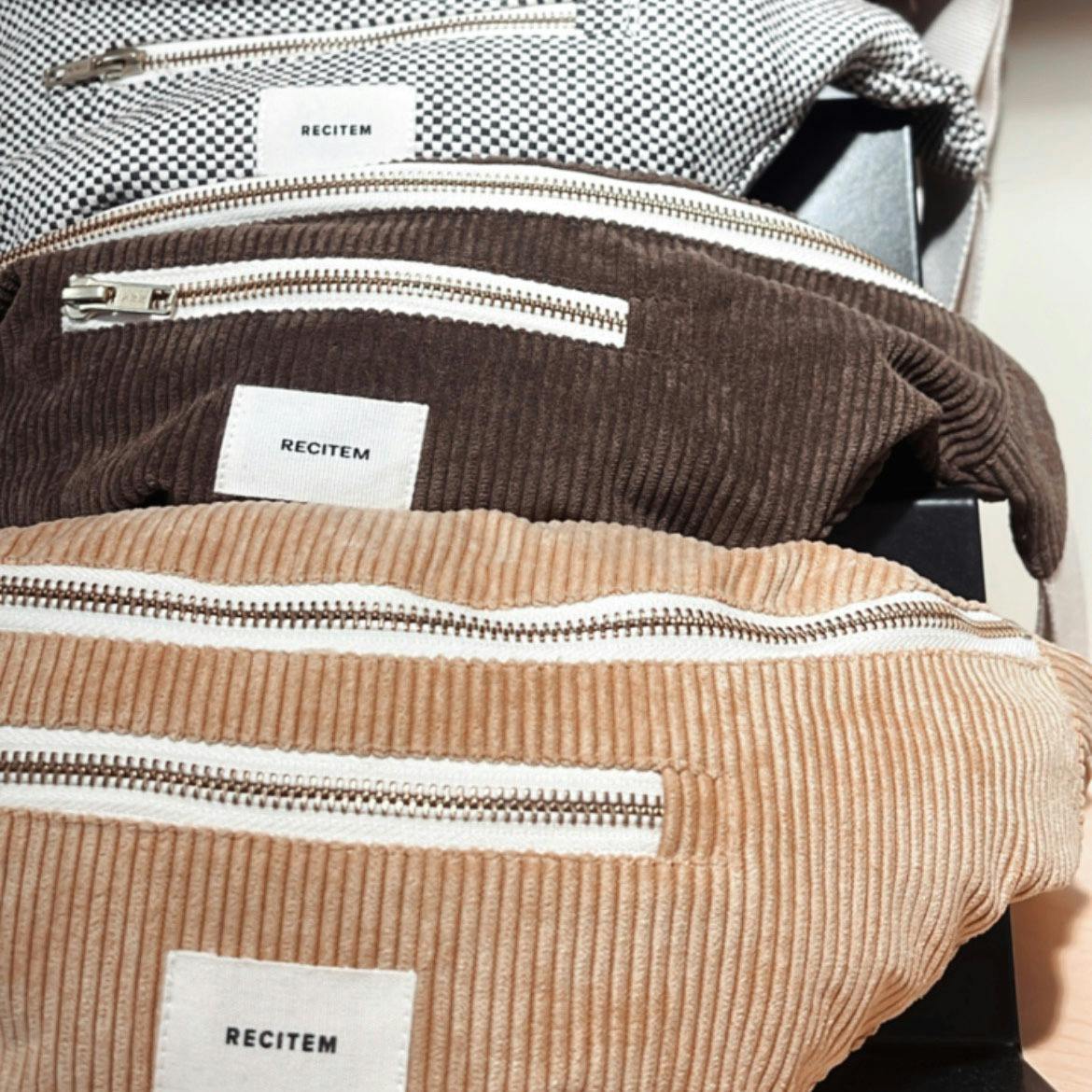 Drei Bauchtaschen aus Cordstoff in Beige, Braun und Schwarz-weiß mit weißen Reißverschlüssen und Etiketten mit einer Aufschrift.