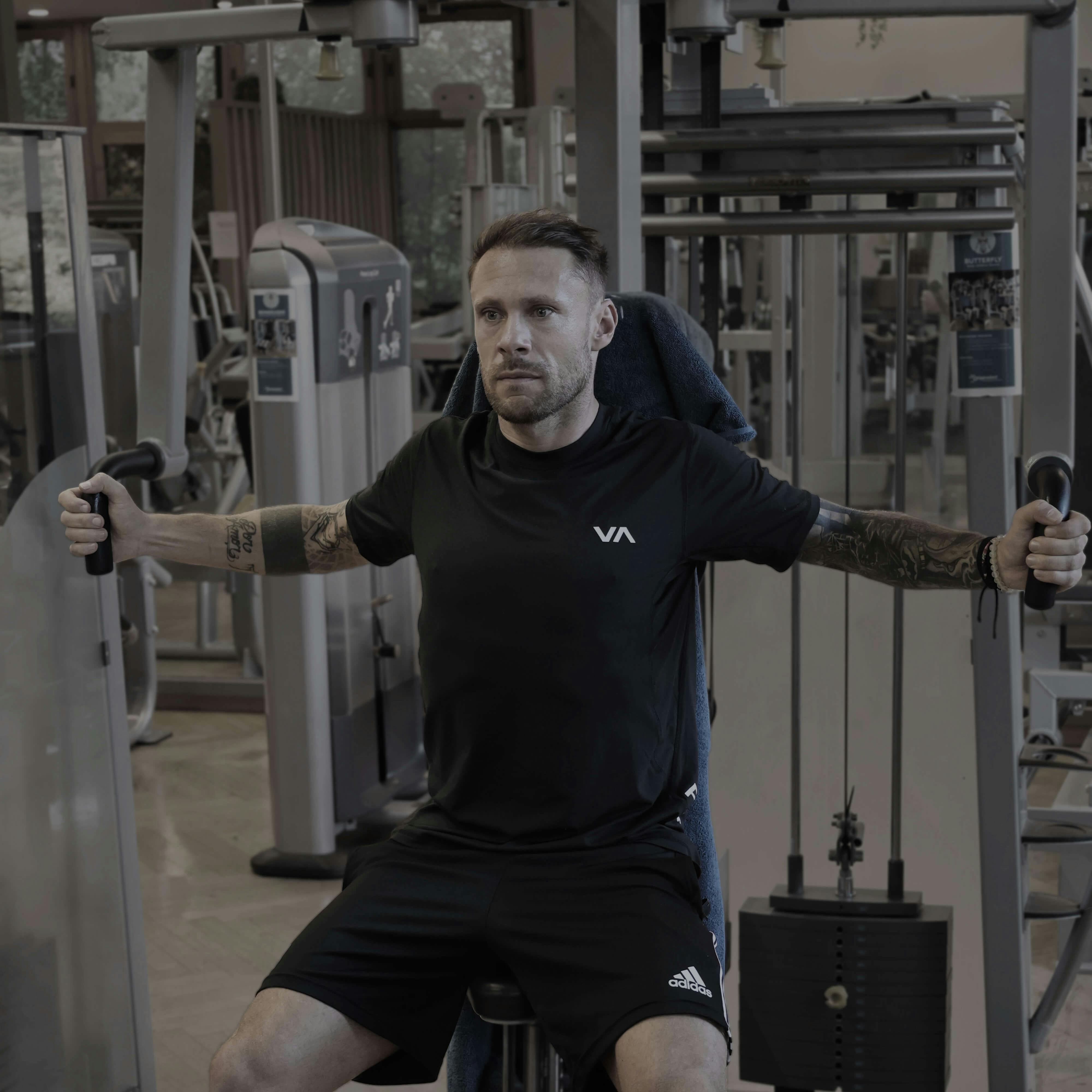 Mann sitzt an Fitnessgerät und führt Butterfly-Übung mit ausgestreckten Armen aus