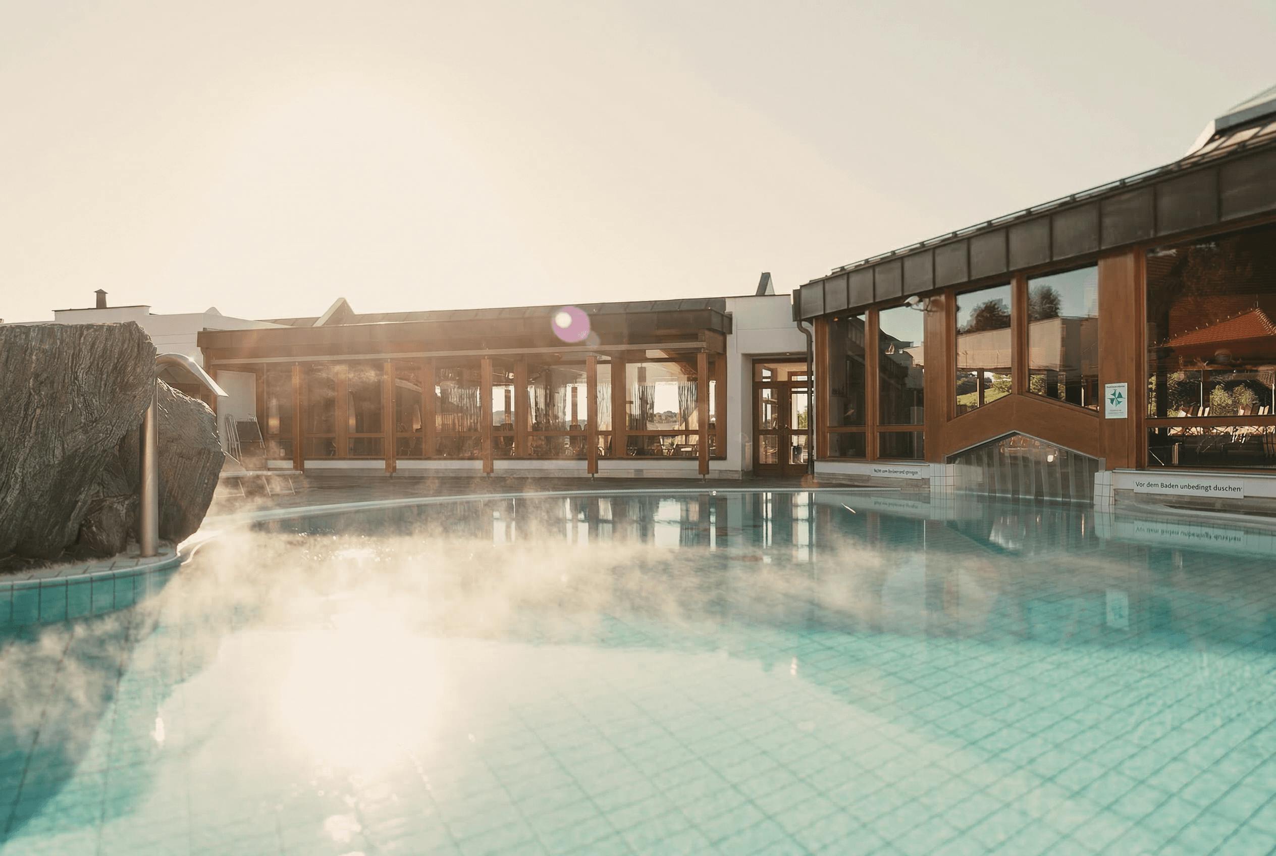 Dampfender Außenpool vor einem Gebäude mit großen Glasfenstern und Holzrahmen bei Sonnenlicht