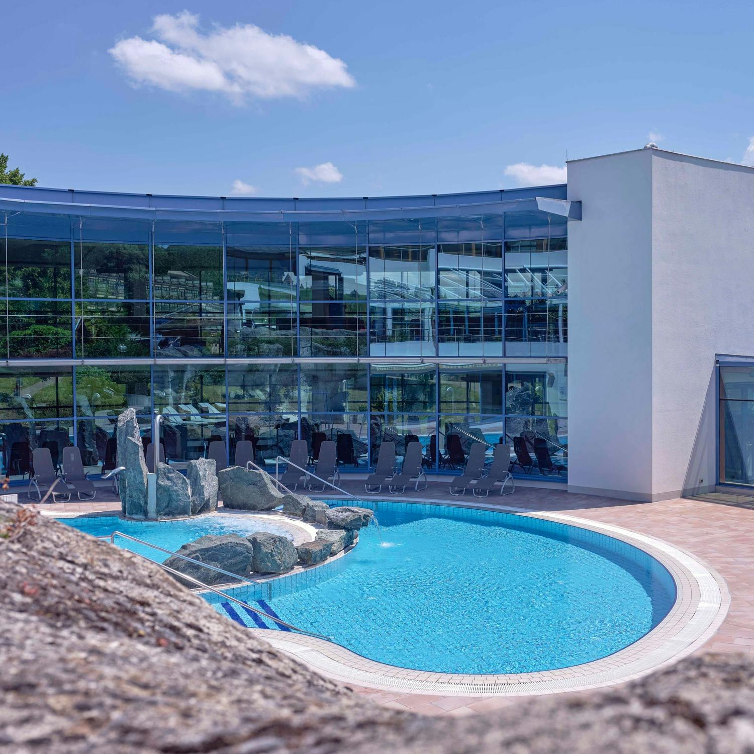 Outdoor pool with rock island in front of a modern building with large glass facade under blue sky
