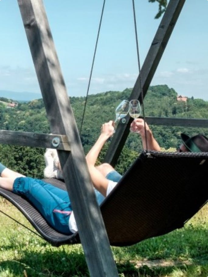 Zwei Menschen liegen in einer Schaukel, blicken auf die Hügel der Steiermark und stoßen mit Gläsern an.
