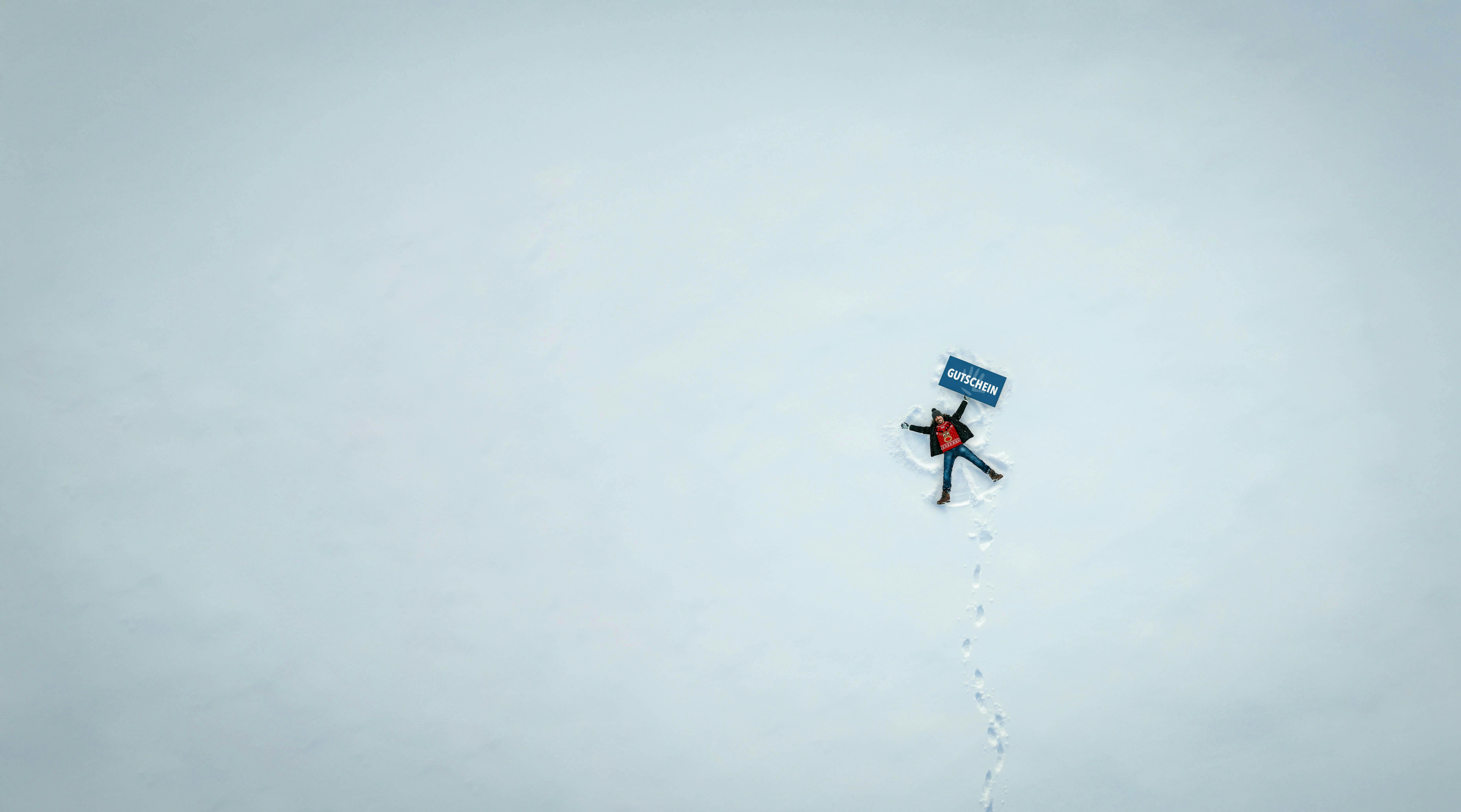 Stani, wearing a red Christmas sweater, is lying on snow-covered ground and holding a blue sign with white lettering that says “Gutschein” (voucher).