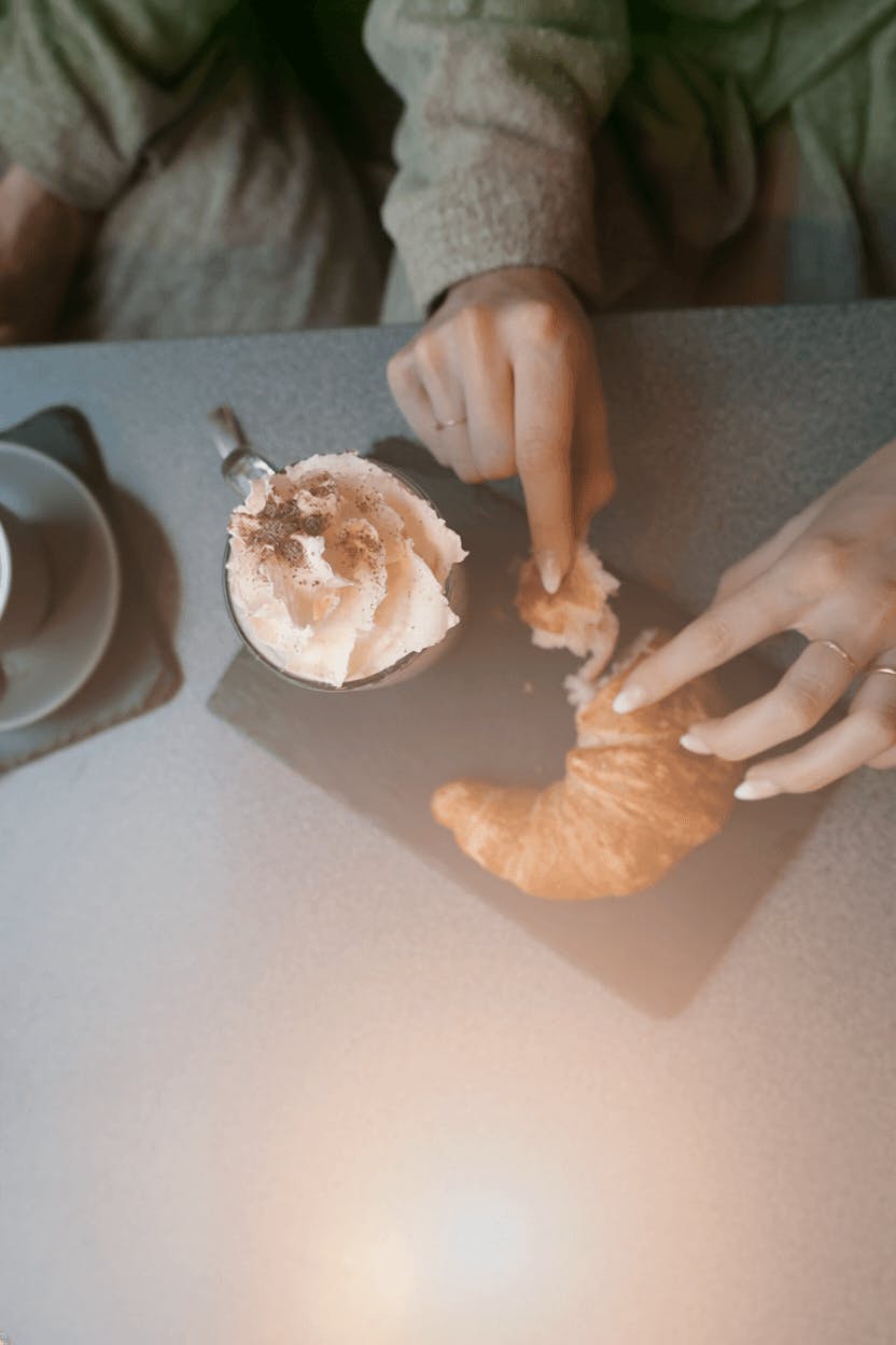 Zwei Hände greifen nach einem Croissant neben einer Tasse Kaffee mit Sahnehaube auf einem Tisch