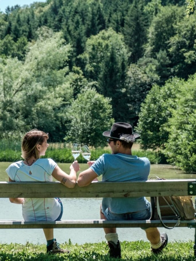 Zwei Menschen die auf einer Bank vor einem See sitzen und mit Gläsern anstoßen.
