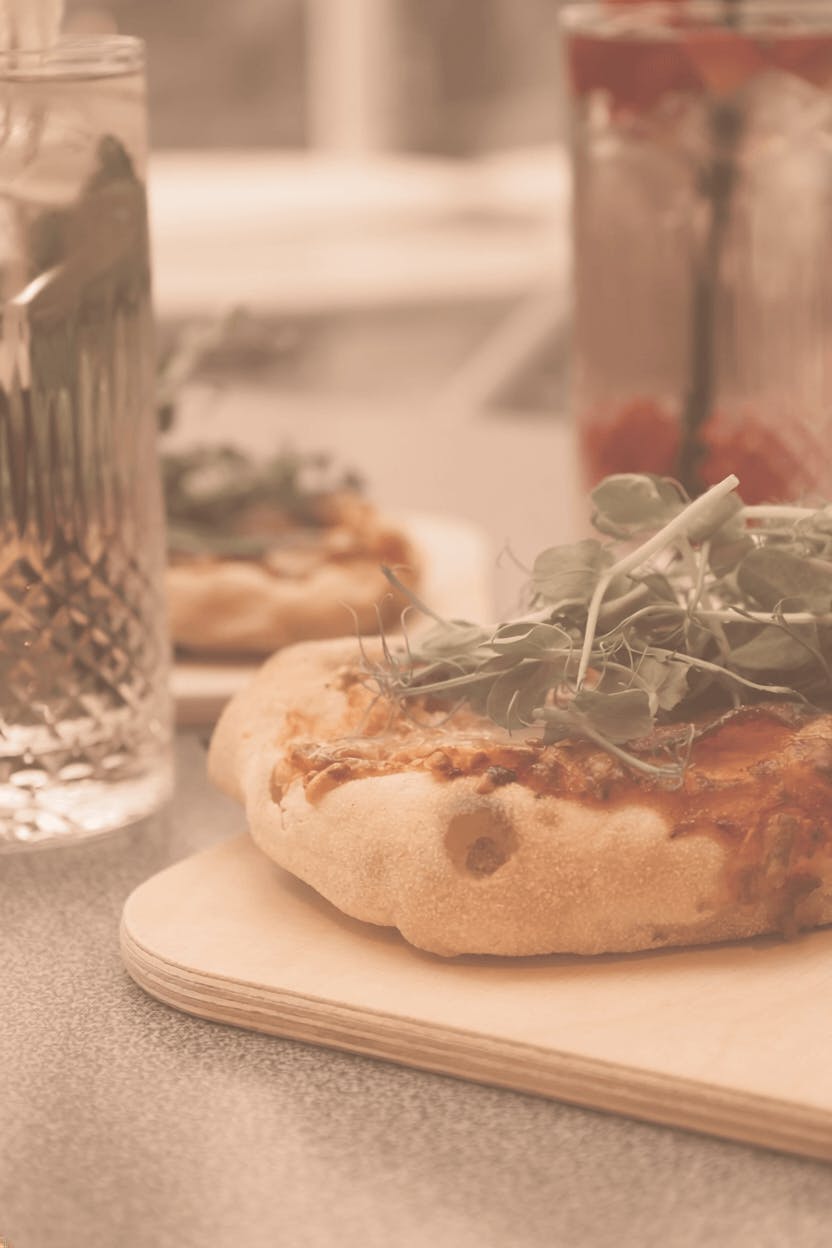 Pizza mit frischem Rucola auf einem Holzbrett neben einem Glas Wasser und einem Glas mit Getränk und Strohhalm