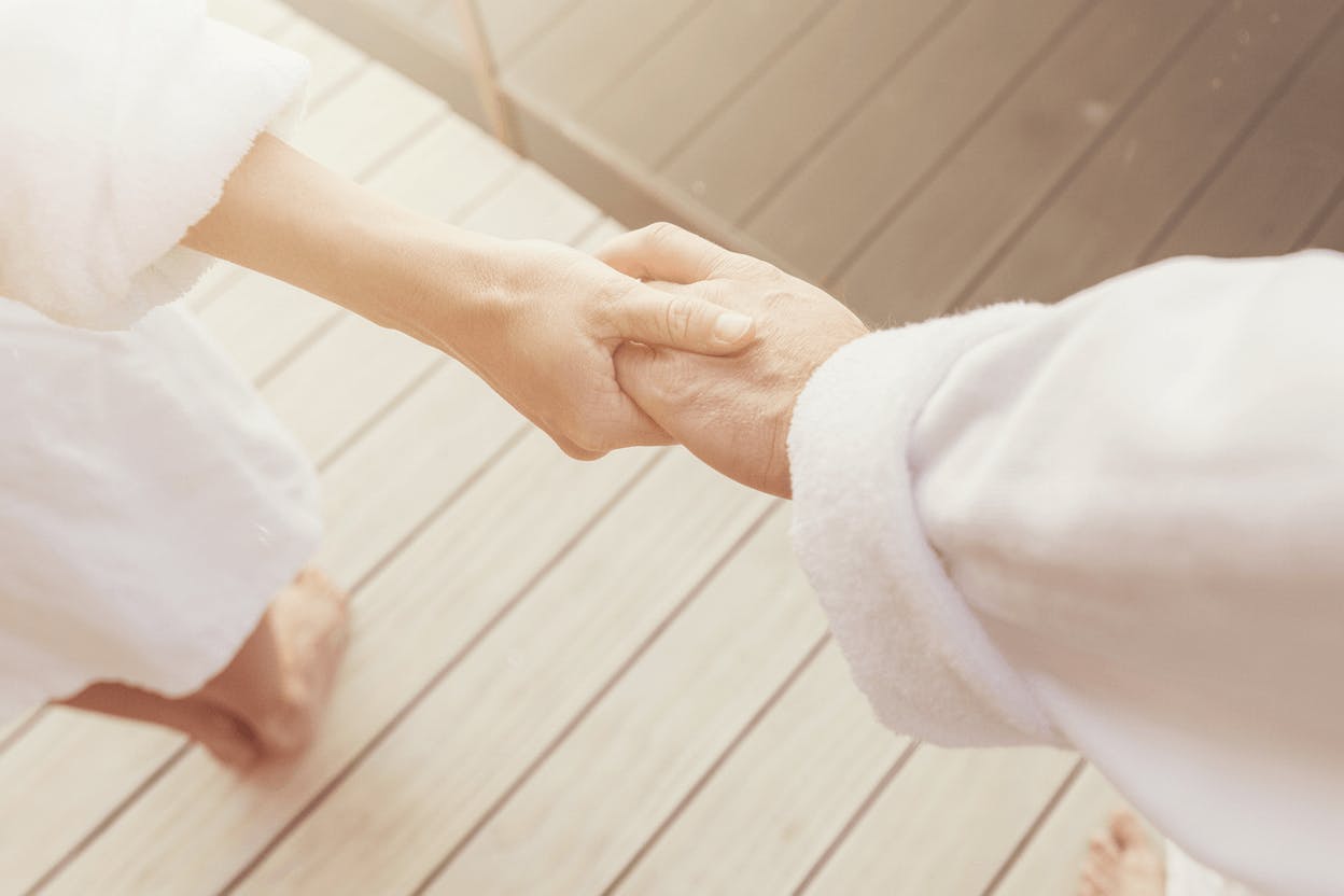 Zwei Personen in Bademänteln halten sich an den Händen stehend auf einem Holzfußboden