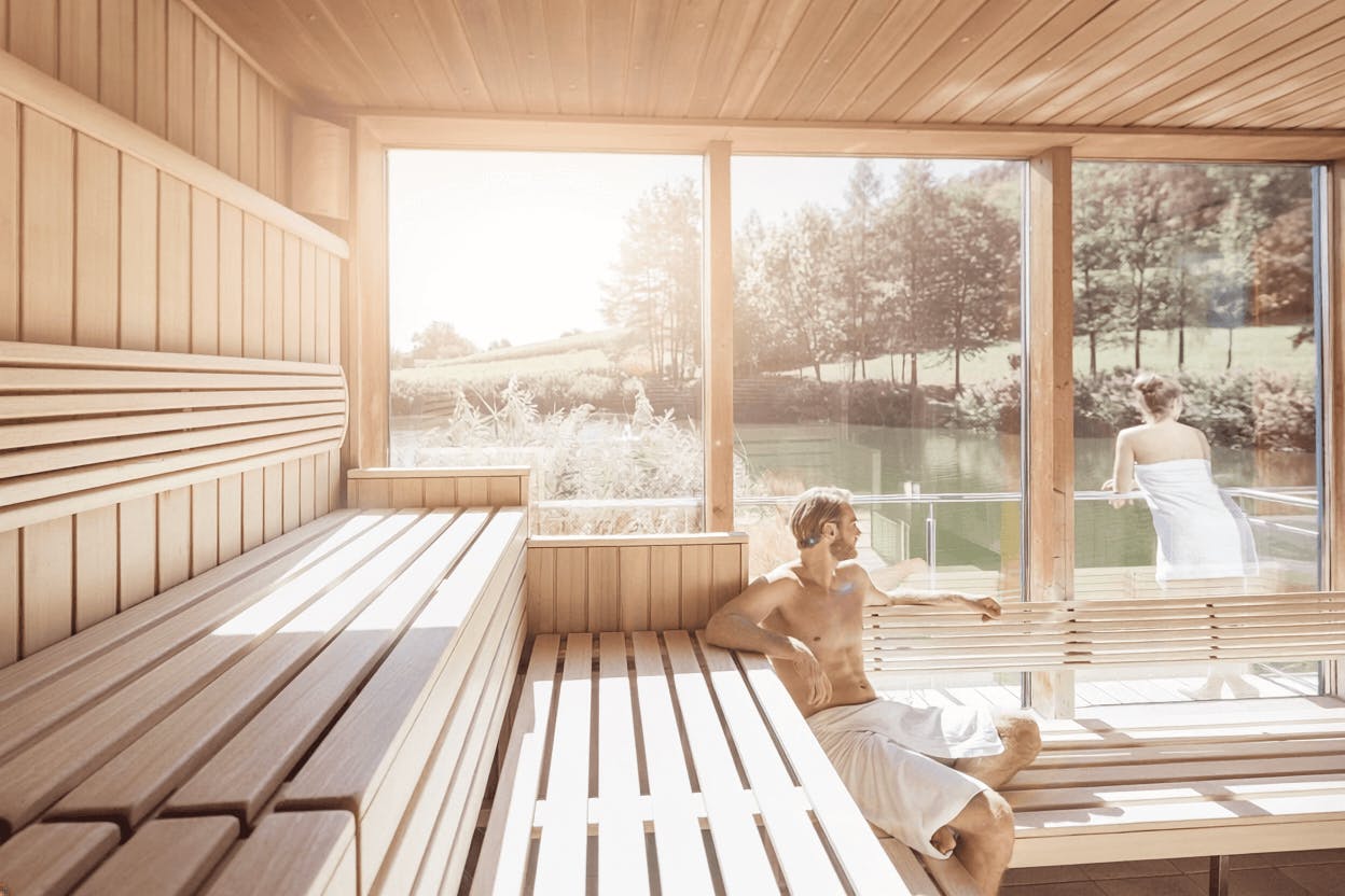 Innenraum einer Sauna mit Holzbänken und großen Fenstern, durch die eine grüne Landschaft sichtbar ist, zwei Personen mit Handtüchern sitzen und stehen auf den Bänken