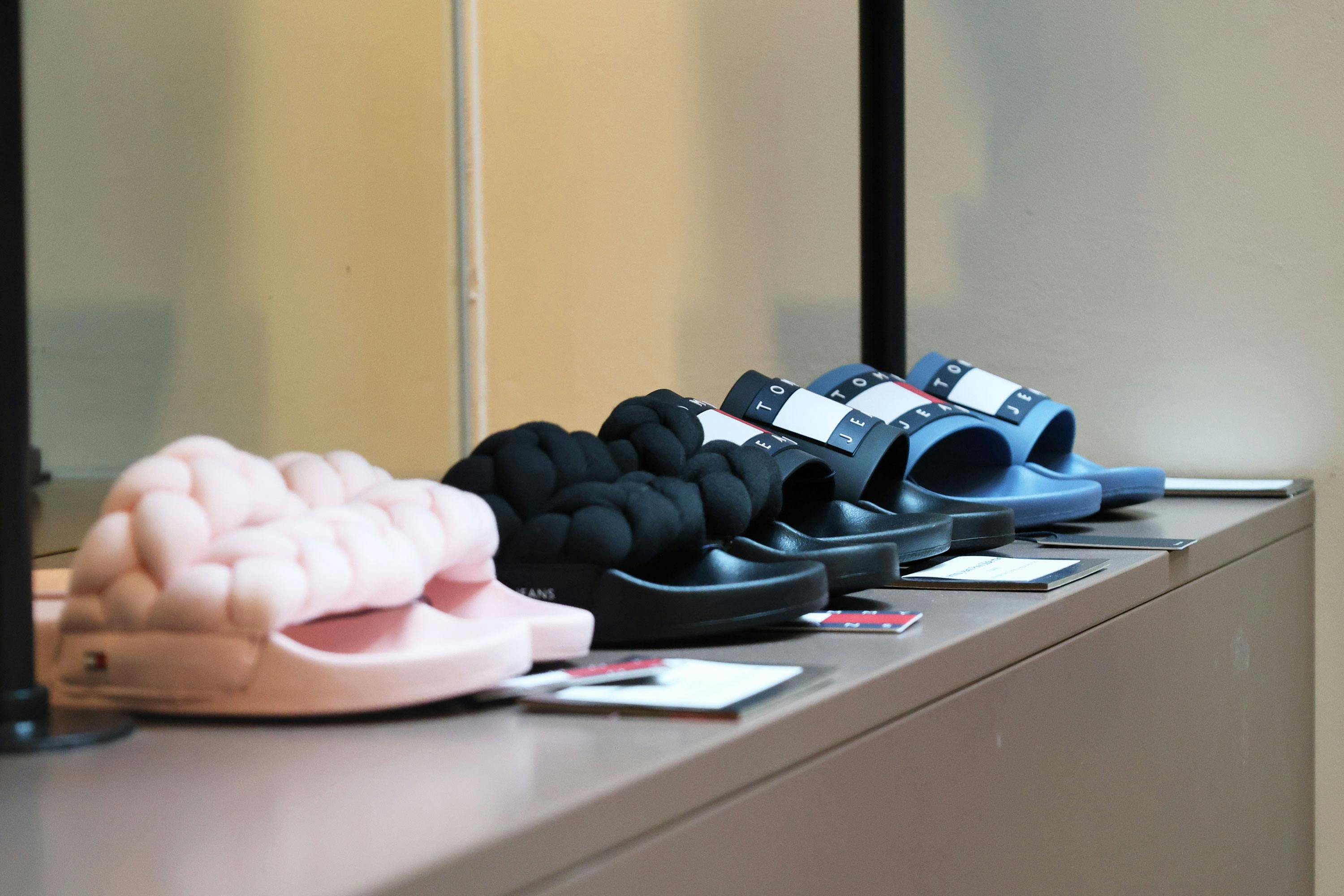 Vier Paar Hausschuhe in Pastellrosa, Schwarz und Blau mit gepolsterten, knotenartigen Riemen auf einer Ablage.