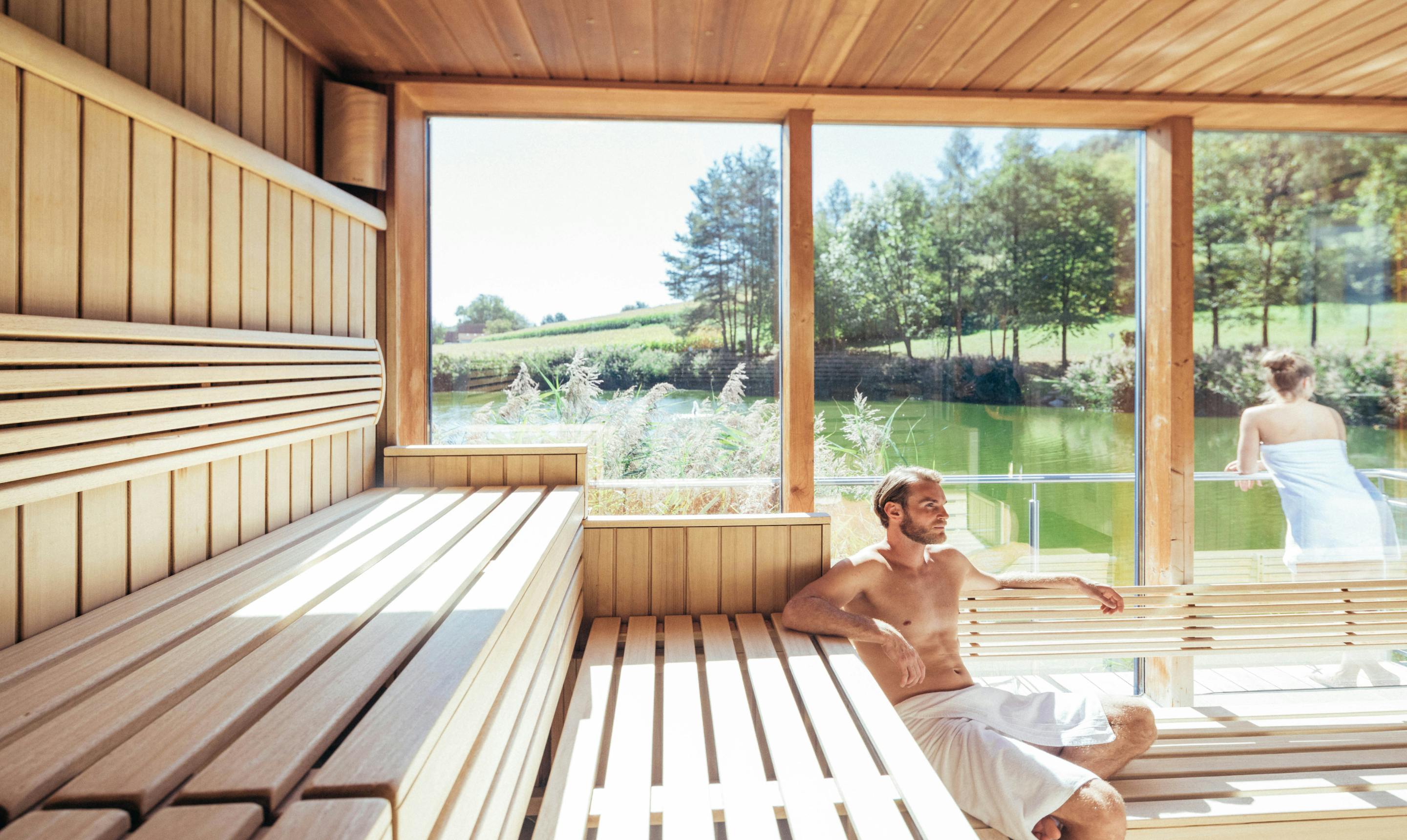 Eine finnische Sauna mit zwei Personen und Ausblick auf einen Teich.