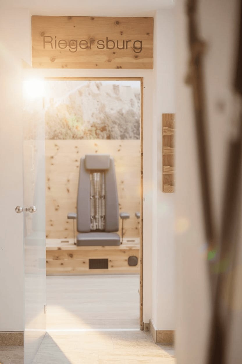 Blick durch eine offene Glastür in einen Raum mit einem grauen Massagestuhl und einer Holztafel mit der Aufschrift 'Riegersburg'