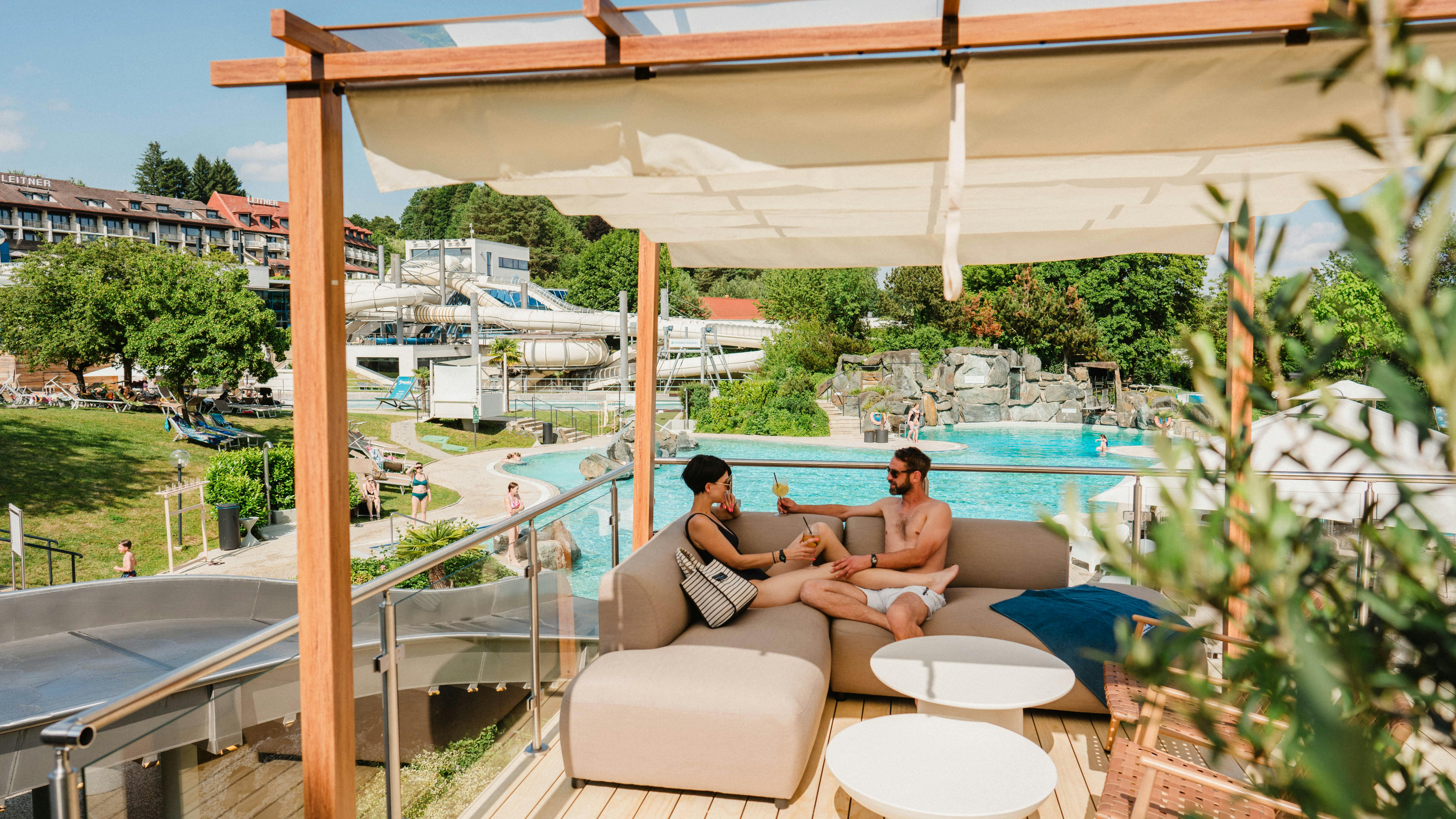 Couple relaxing on a lounge sofa on the terrace with a view of the pool and water slides in the outdoor area of the Loipersdorf thermal spa.