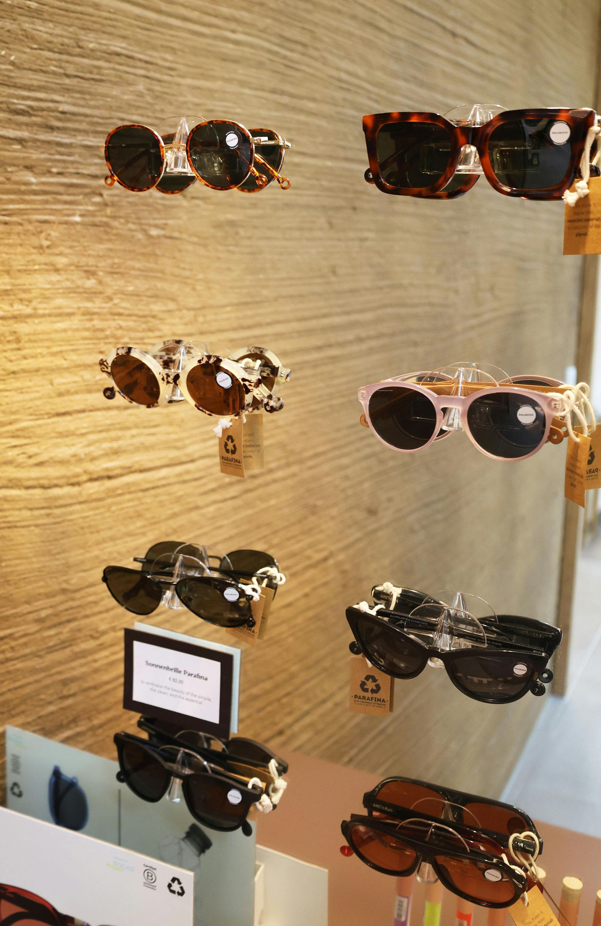 Mehrere Sonnenbrillen mit unterschiedlichen Rahmenfarben und -formen, darunter braun, schwarz und transparent, an einer vertikalen Halterung vor einer strukturierten Wand.