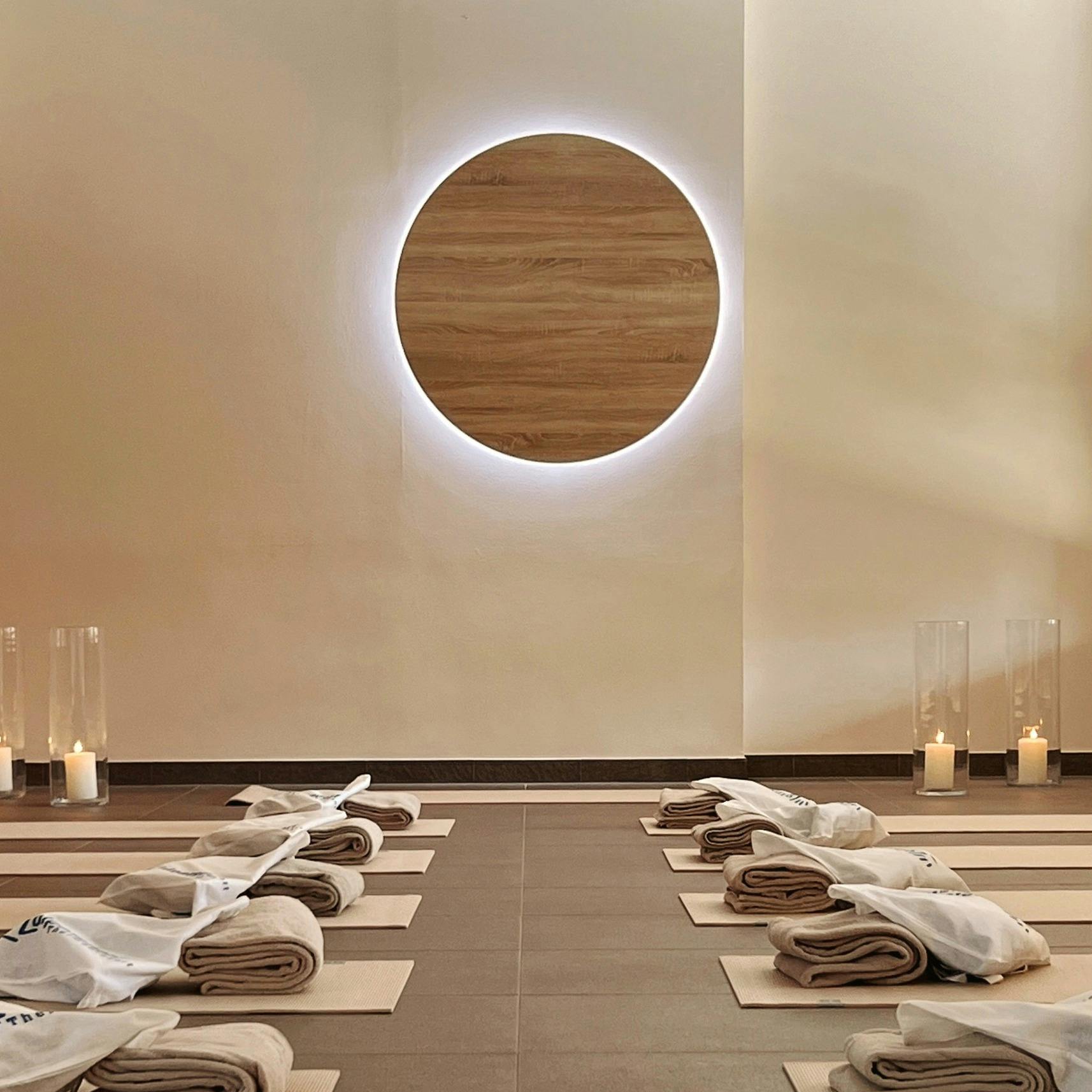 Symmetrically arranged yoga mats with folded blankets and towels in front of an illuminated, round wooden decorative panel on the wall.