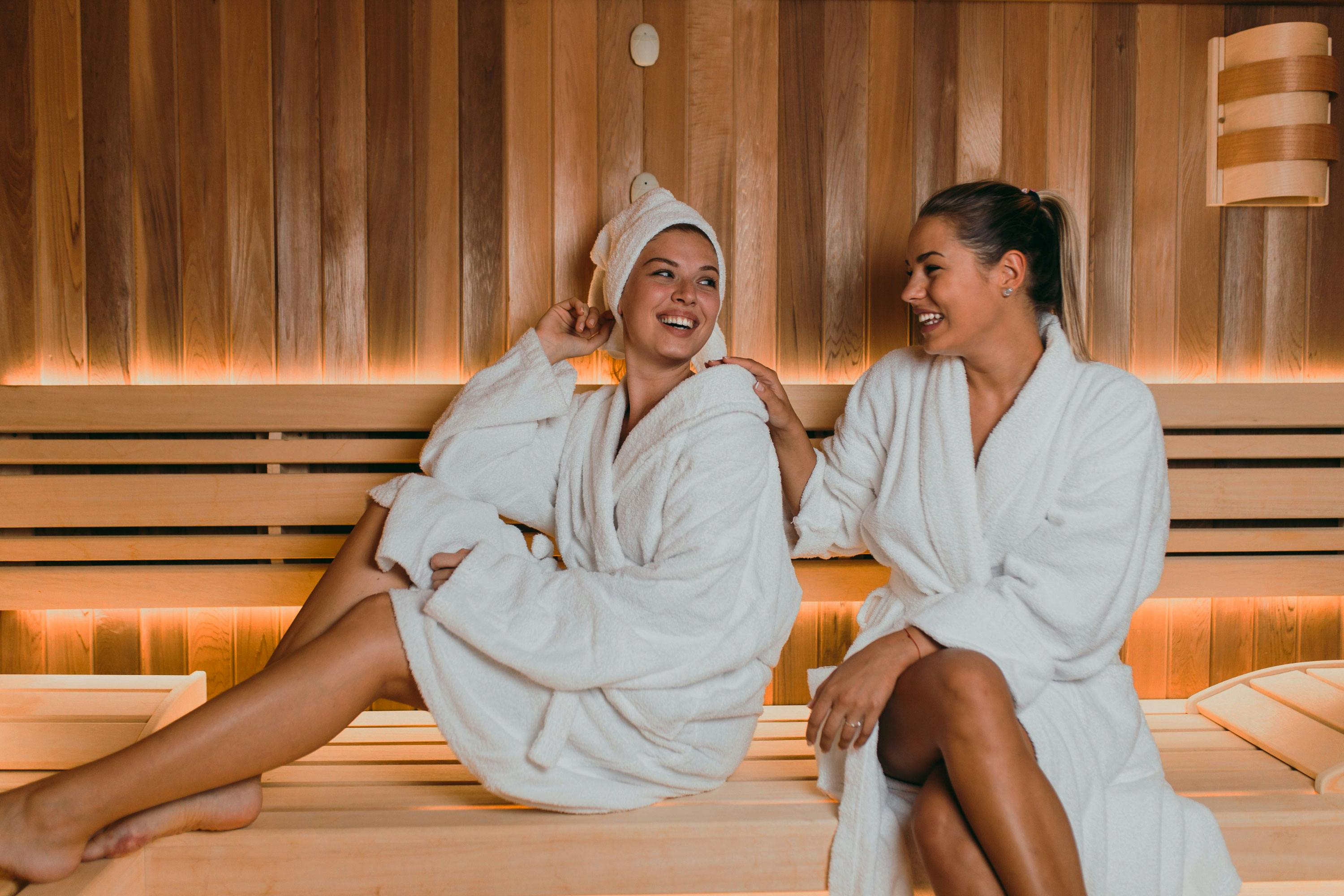 Two women in white bathrobes sit relaxed on a wooden bench in a sauna with warm lighting.