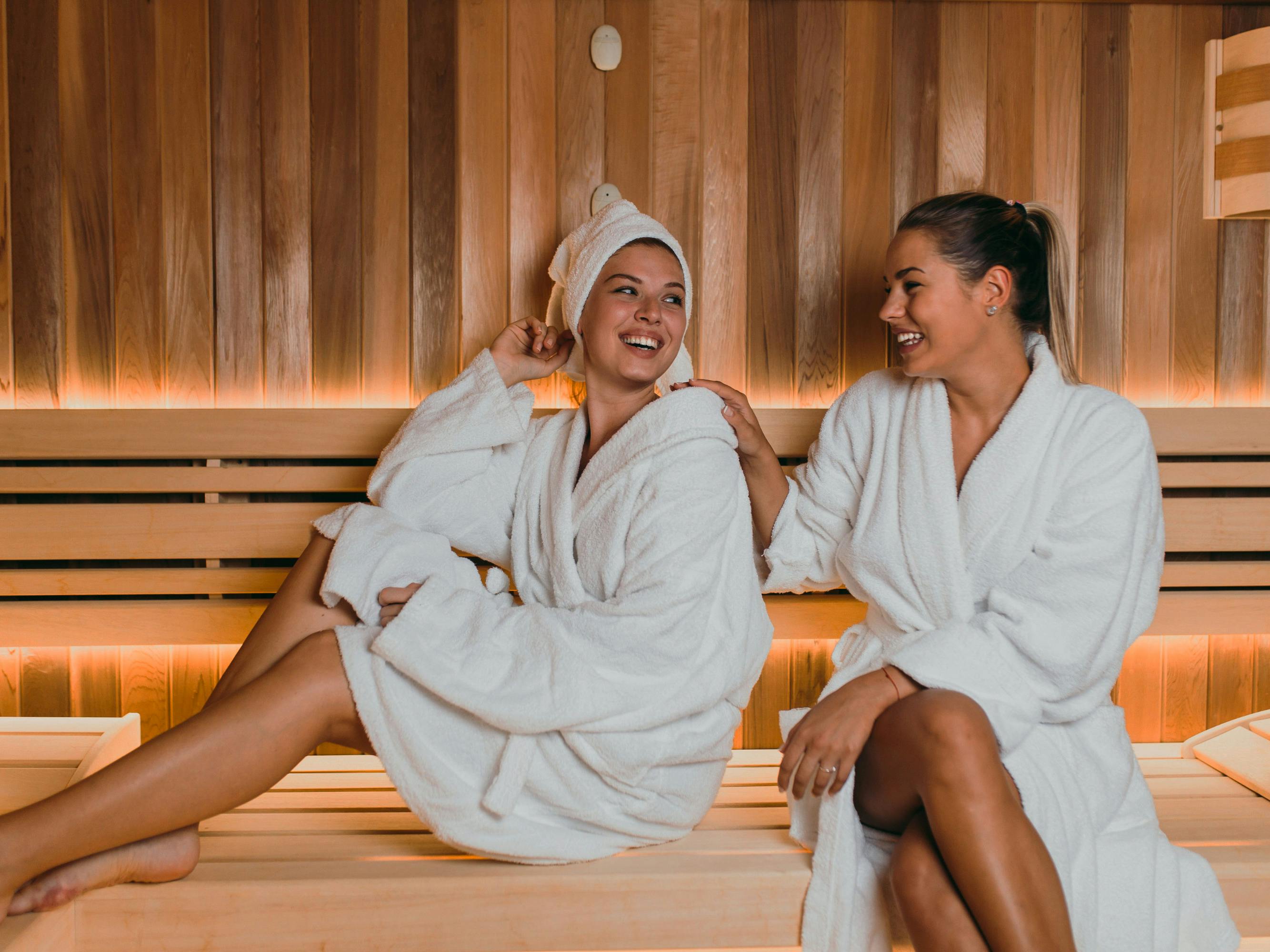 Two women in white bathrobes sit relaxed on a wooden bench in a sauna with warm lighting.