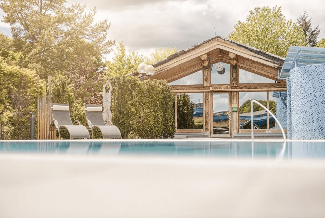 Außenpool mit Liegestühlen und Holzhütte im Hintergrund, umgeben von Bäumen