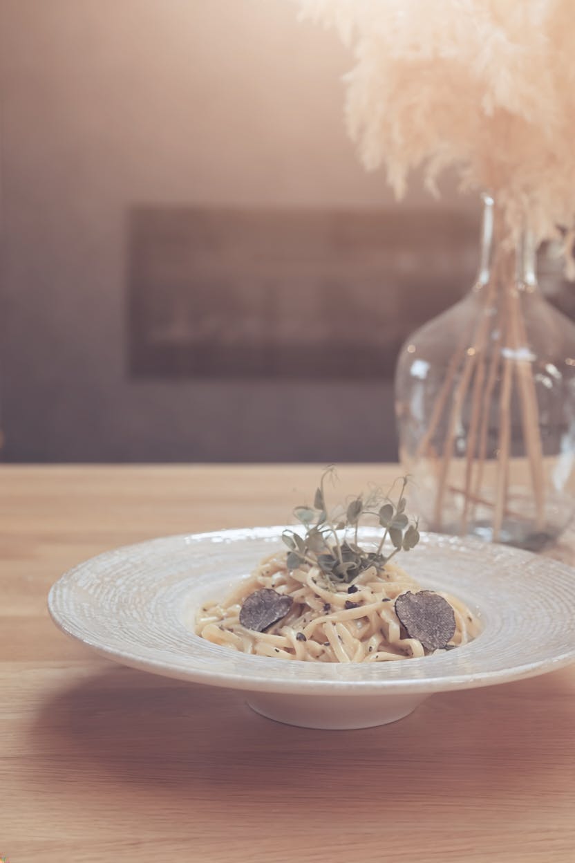 Plate of pasta garnished with truffle slices and fresh herbs on a wooden table in front of a vase of dried flowers.